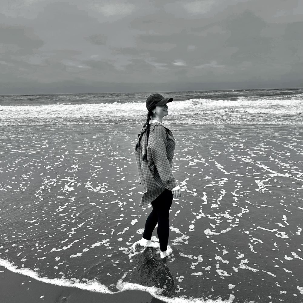 Jaden in a cap and plaid shirt, barefoot in the surf on a cloudy beach, smiling toward the sky