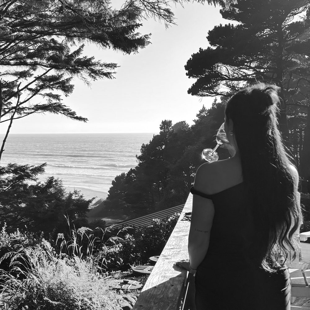 Jaden from behind, holding a glass on a deck above pine trees with the ocean in the distance